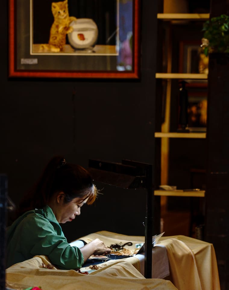 A woman in a green shirt bends over her embroidery frame in a dark workshop, a framed picture of a kitten on the wall above her — Jim Kerns