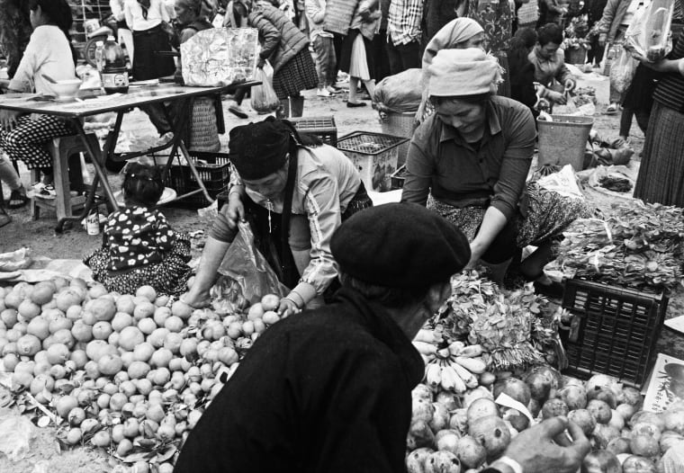 Buyers and sellers lean over piles of pomelo, bananas and green plums at the morning market, a man in a beret in the foreground — Dennis and Patrick Weinert