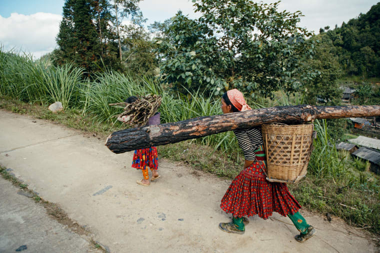 A woman carries a whole timber beam on her shoulder along a concrete path, a woven basket on her back and a small child walking ahead with a bundle of sticks — Dennis and Patrick Weinert