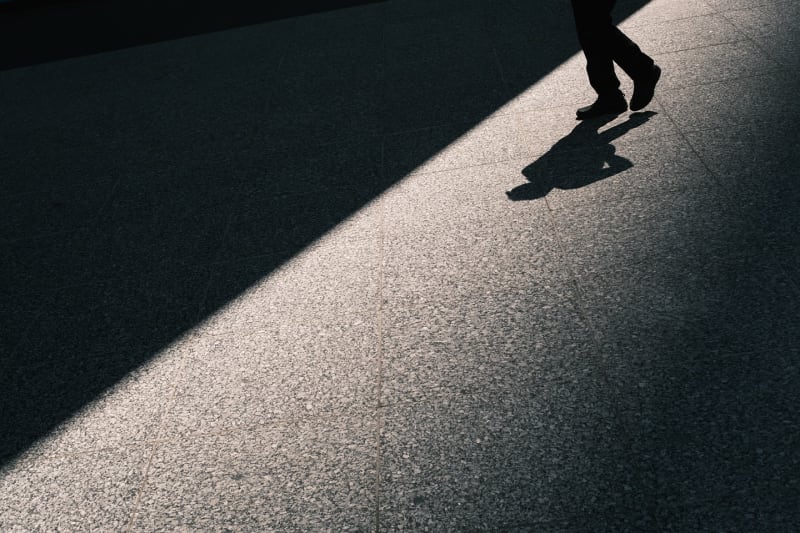 Overhead frame of a lone walker casting a long shadow across the diagonal edge of sunlit pavement