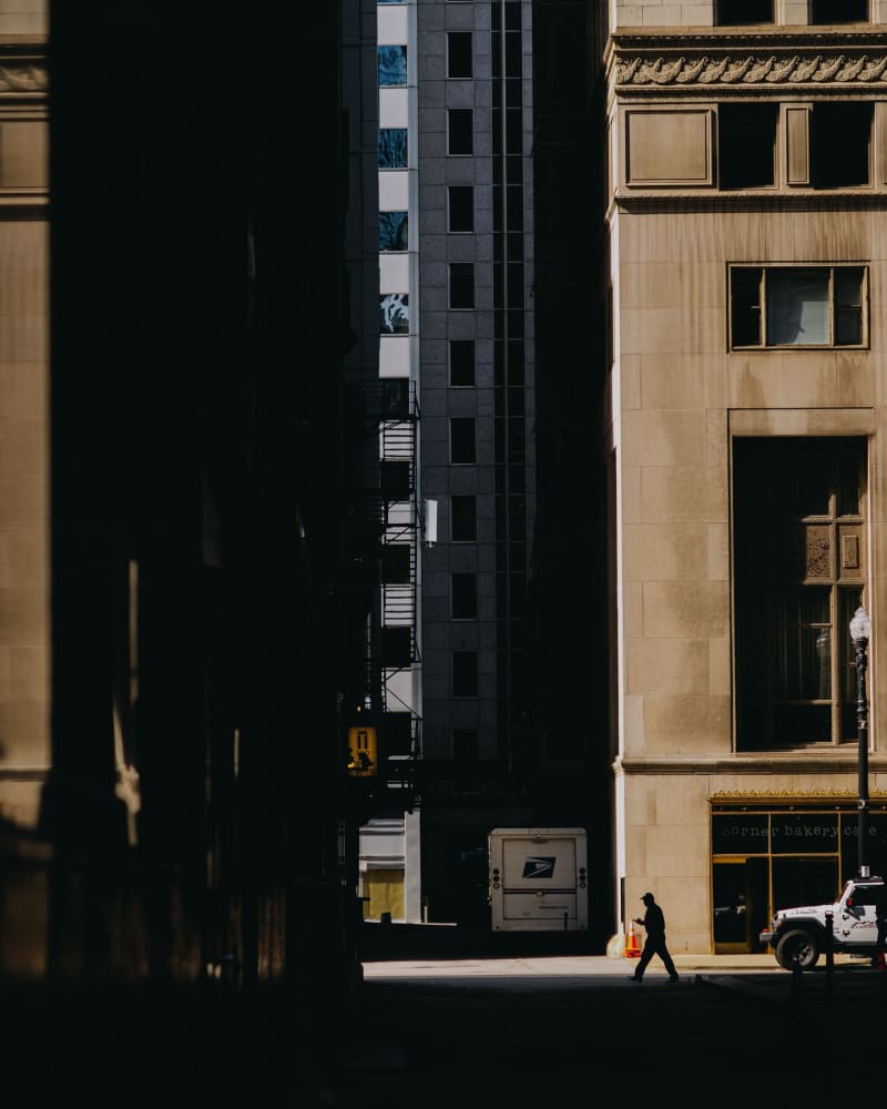 Late light raking across two Loop high-rise facades, a single figure crossing the street far below