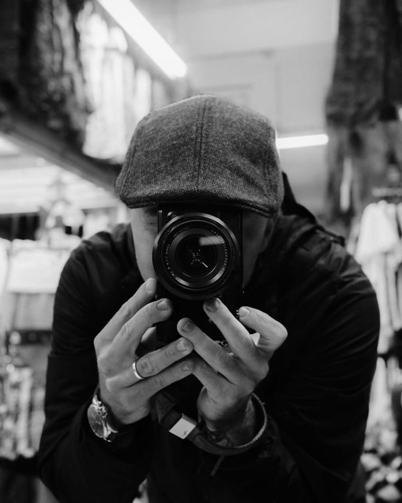 Jack Ross in a flat cap, shooting a black-and-white self-portrait with a rangefinder-style camera raised to his eye