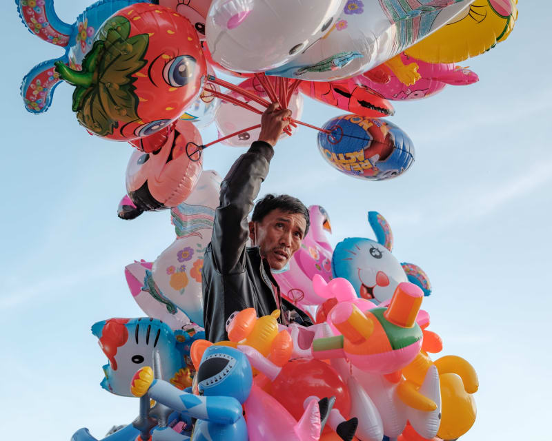 A balloon seller reaches up into a crowded bunch of brightly coloured foil balloons against a pale sky