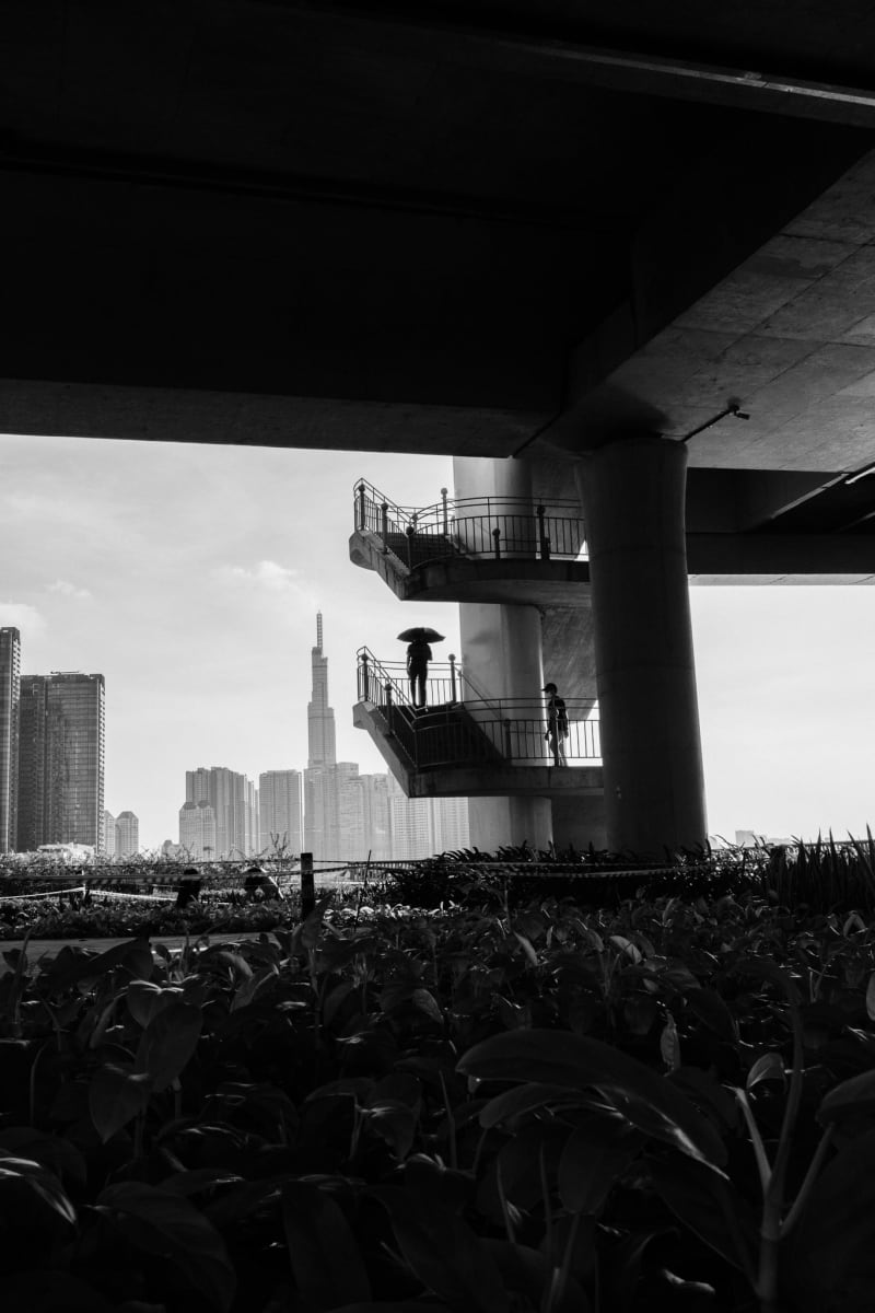 Black-and-white view from beneath a flyover — switchback stairs, a lone figure with an umbrella, and the Saigon skyline beyond