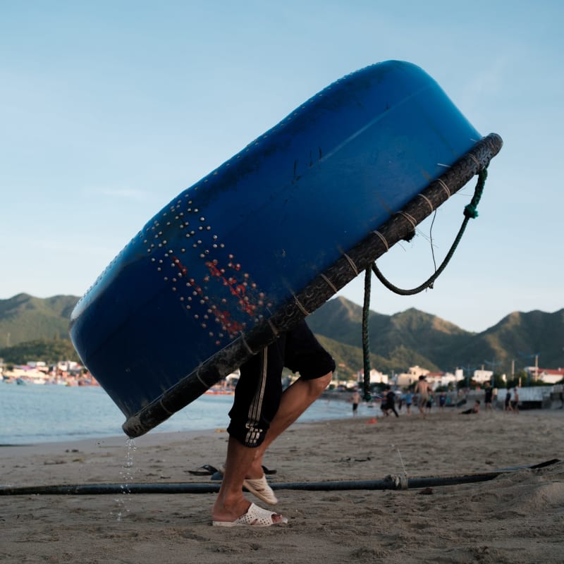 A fisherman carries a blue round coracle over his head across the sand, dripping water