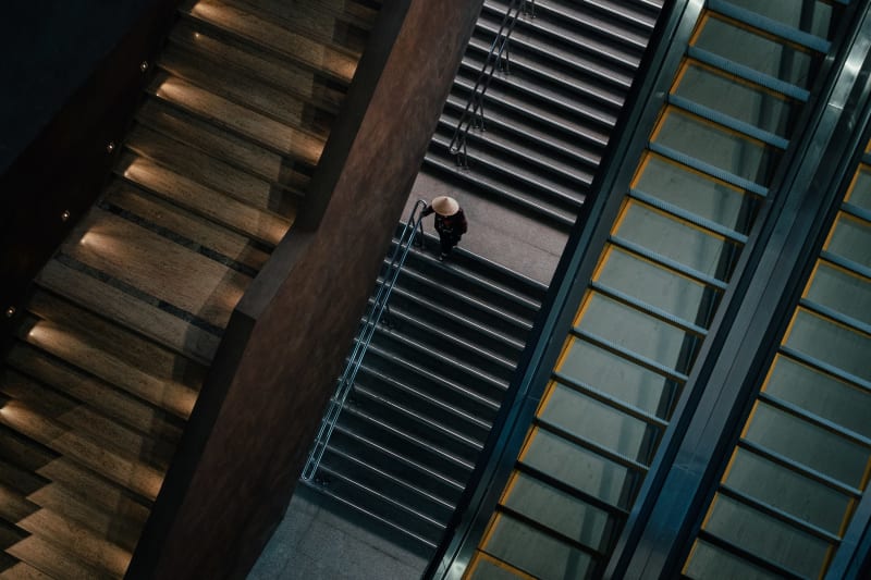 Overhead frame of stairs and escalators converging, a small figure in a conical hat crossing the landing between them