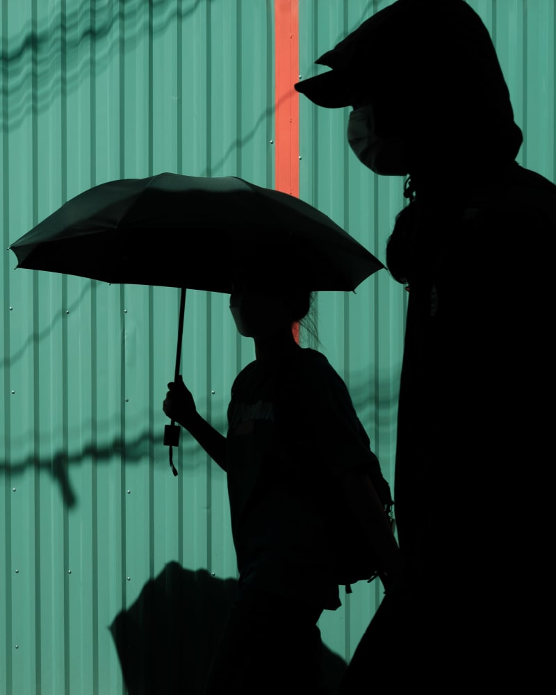 Two masked figures in silhouette against a teal corrugated wall, one holding a black umbrella