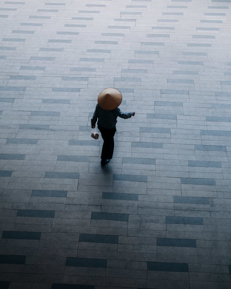 Overhead frame of a lone figure in a conical hat crossing a wide grey tiled plaza