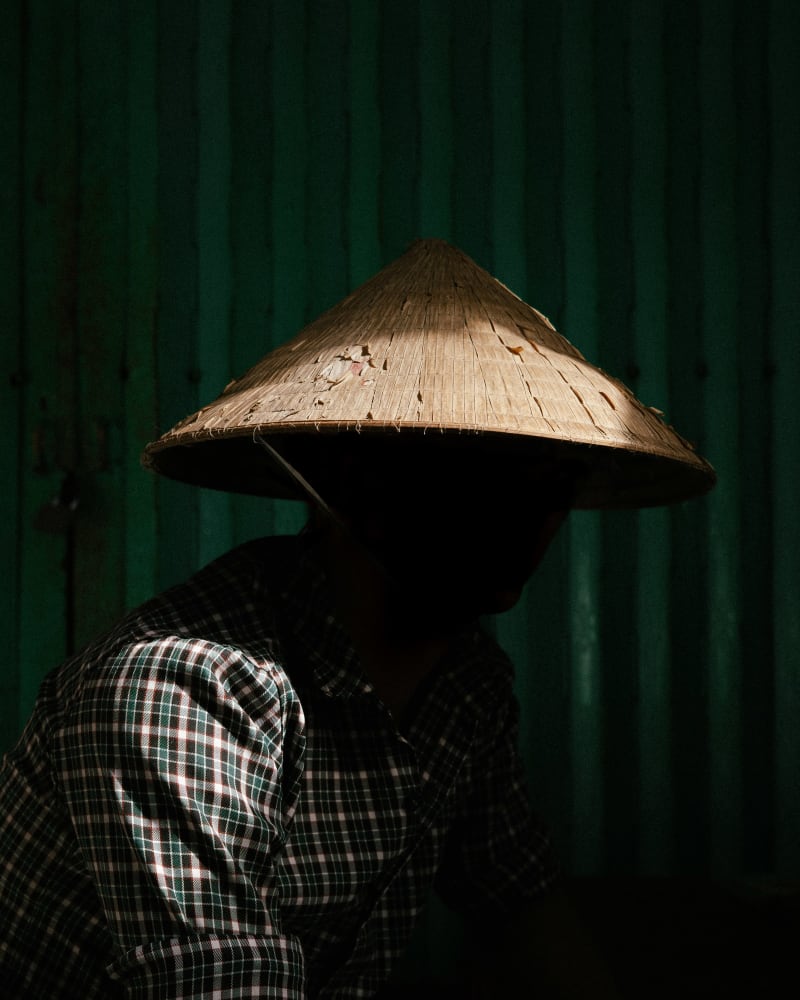 A worker in a nón lá and checked shirt lit by a single shaft of light against a dark green wall