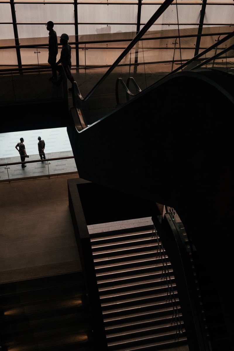Figures silhouetted on an upper walkway against bright dusk light, dark stair geometry cutting across below