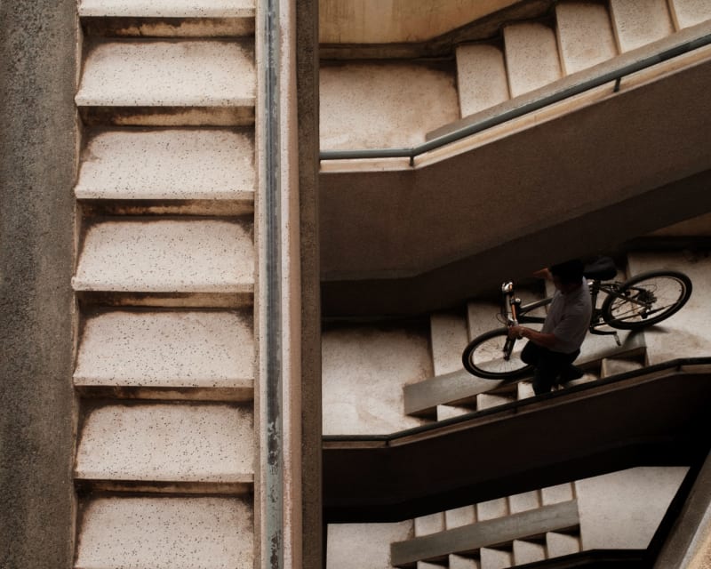 Seen from above, a man carries a bicycle down a concrete apartment stairwell