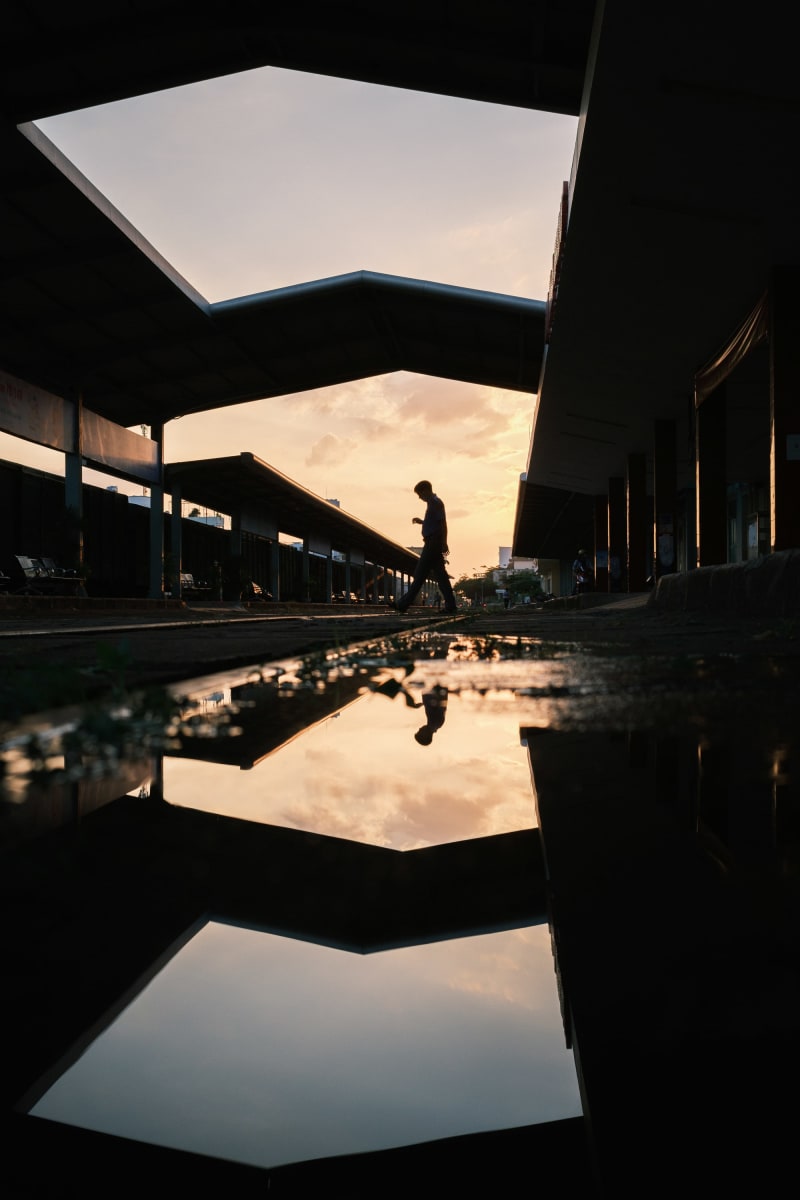 A man walks between station canopies at sunset, mirrored in a still puddle in the foreground