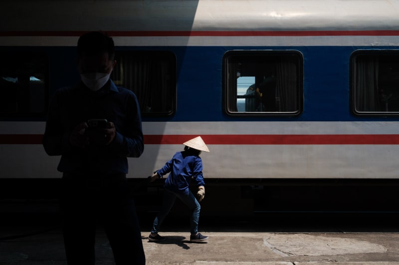 A woman in a conical hat crosses a sunlit strip on a train platform while a silhouetted man checks his phone