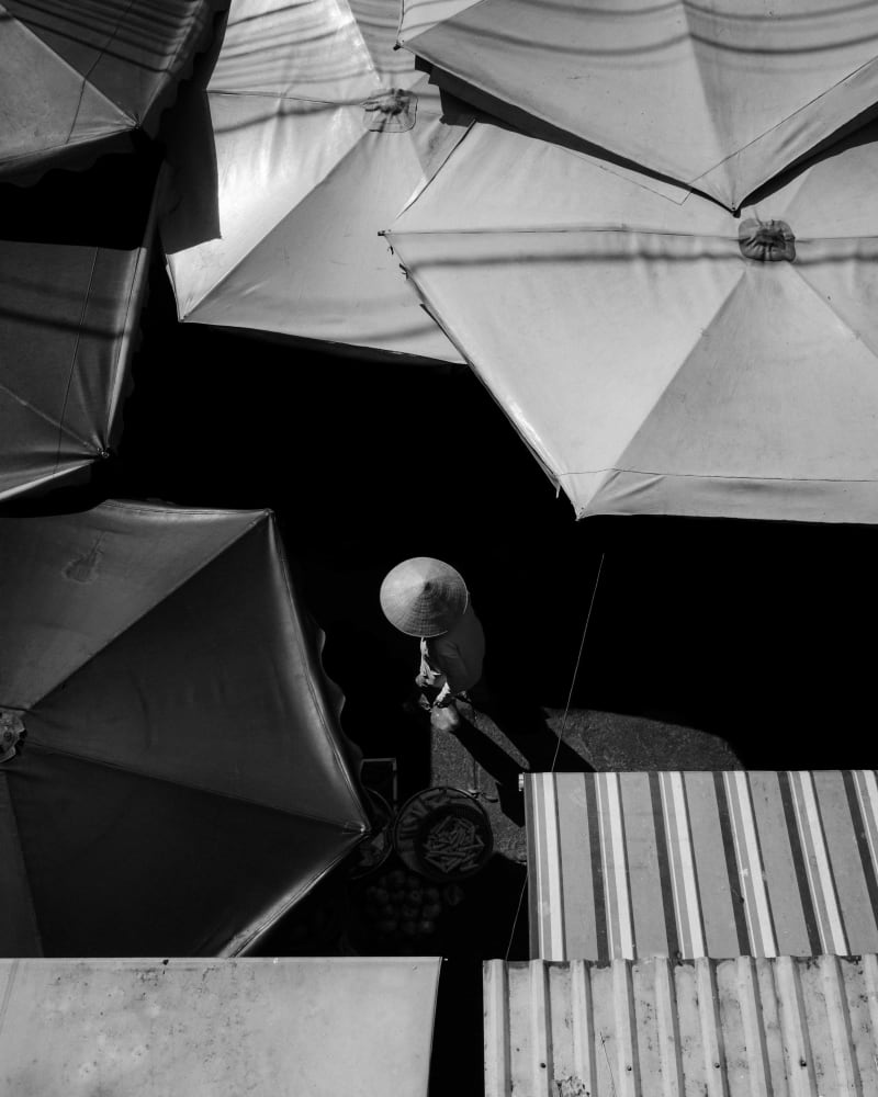 Black-and-white overhead view of market umbrellas, a vendor in a conical hat in the gap between them