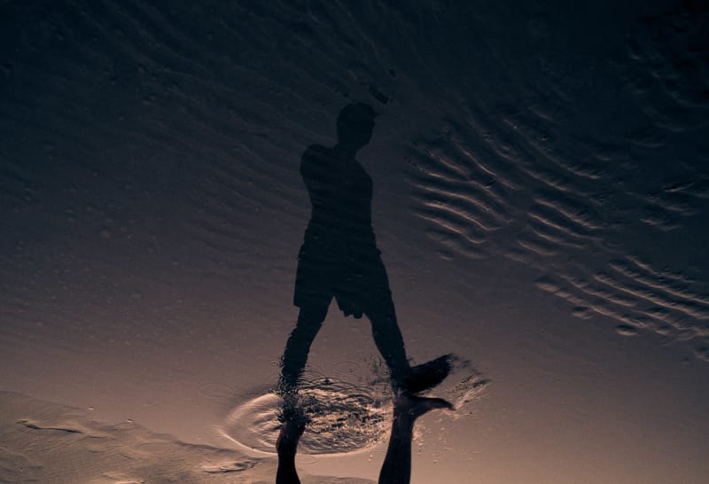 A figure's mirrored reflection in a thin sheet of water on rippled sand, feet breaking the surface