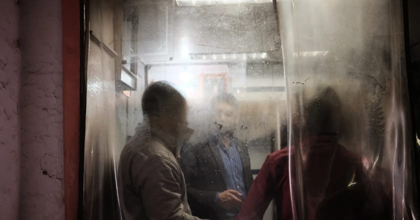 Three men stand at a night food stall, their figures softened by a scratched sheet of hanging plastic between them and the camera — Luke Neville
