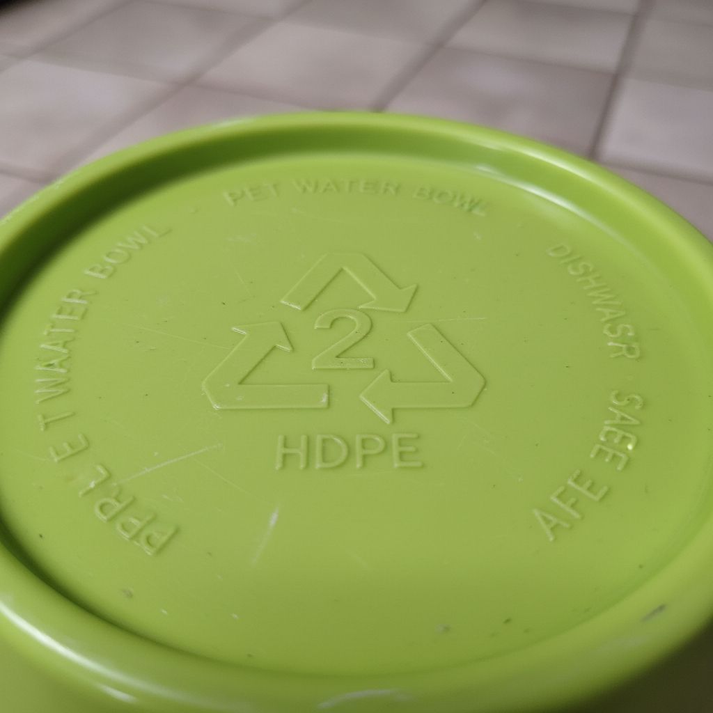 A macro, photorealistic shot of the bottom of a lime green HDPE pet water bowl. The focus is sharp on the molded recycling symbol, clearly showing the number '2' inside the chasing arrows triangle. The plastic texture is detailed. 4k.