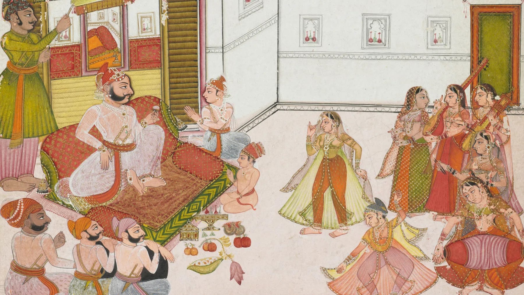 An opaque watercolor and ink painting on paper depicts a lively courtly gathering within a white-walled terrace or open courtyard. On the left, a bearded ruler with a prominent mustache sits cross-legged on a large, low, patterned cushion supported by red bolsters. He is dressed in a translucent white jama robe, an ornate red and gold turban, and fine jewelry as he looks toward a performance. Behind him, a standing attendant in a green gown holds a fly-whisk, while in front of his platform, two young boys sit on the floor, one with hands pressed together in a respectful gesture. In the lower-left corner, three male courtiers in turbans sit on the ground with their backs partially turned to the viewer.The right side of the painting centers on the entertainment, where two female dancers perform; one stands elegantly in a yellow and green dress, while another spins dynamically in a pink outfit. They are accompanied by a tight cluster of five female musicians on the far right playing traditional instruments, including a stringed tanpura and a small dholak hand drum. The architectural setting features a white wall adorned with small rectangular niches containing red decorative vessels, alongside an open green doorway on the far right. In the foreground between the ruler and the performers, a careful arrangement of offerings is spread across the floor, featuring small golden containers, a lotus bud, and fresh fruits like oranges and pomegranates.