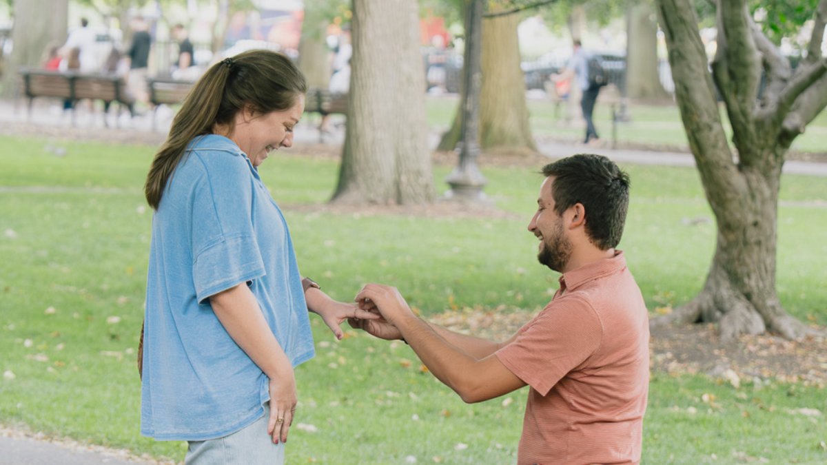 Couple celebrating engagement after surprise proposal at Boston Public Garden with autumn foliage and iconic footbridge in background
