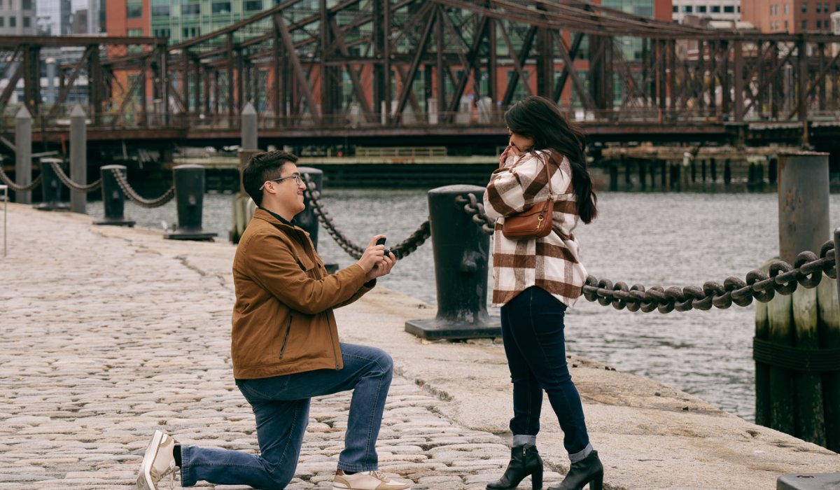 Newly engaged couple celebrating proposal at Boston Seaport waterfront with harbor views and city skyline at golden hour