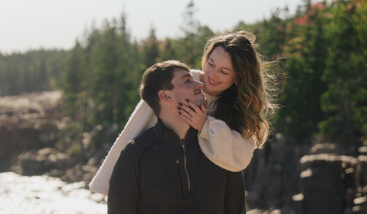 Engaged couple standing on rocky Maine coastline at Acadia Ocean Path during golden hour with waves crashing on granite cliffs