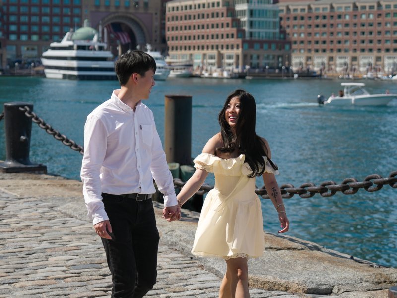 Couple laughing together during engagement session at Boston Seaport with city skyline reflecting in harbor at dusk