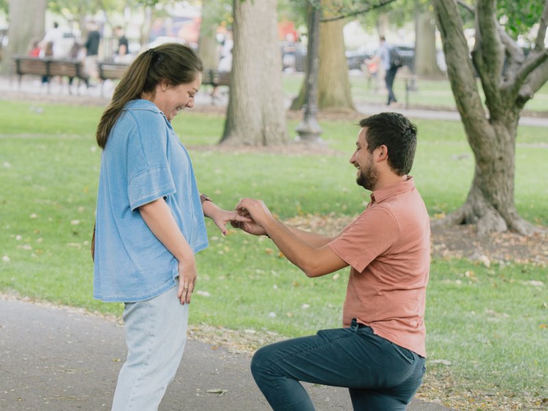 Man proposing to girlfriend on one knee at center of Boston Public Garden footbridge during golden hour with perfect symmetry