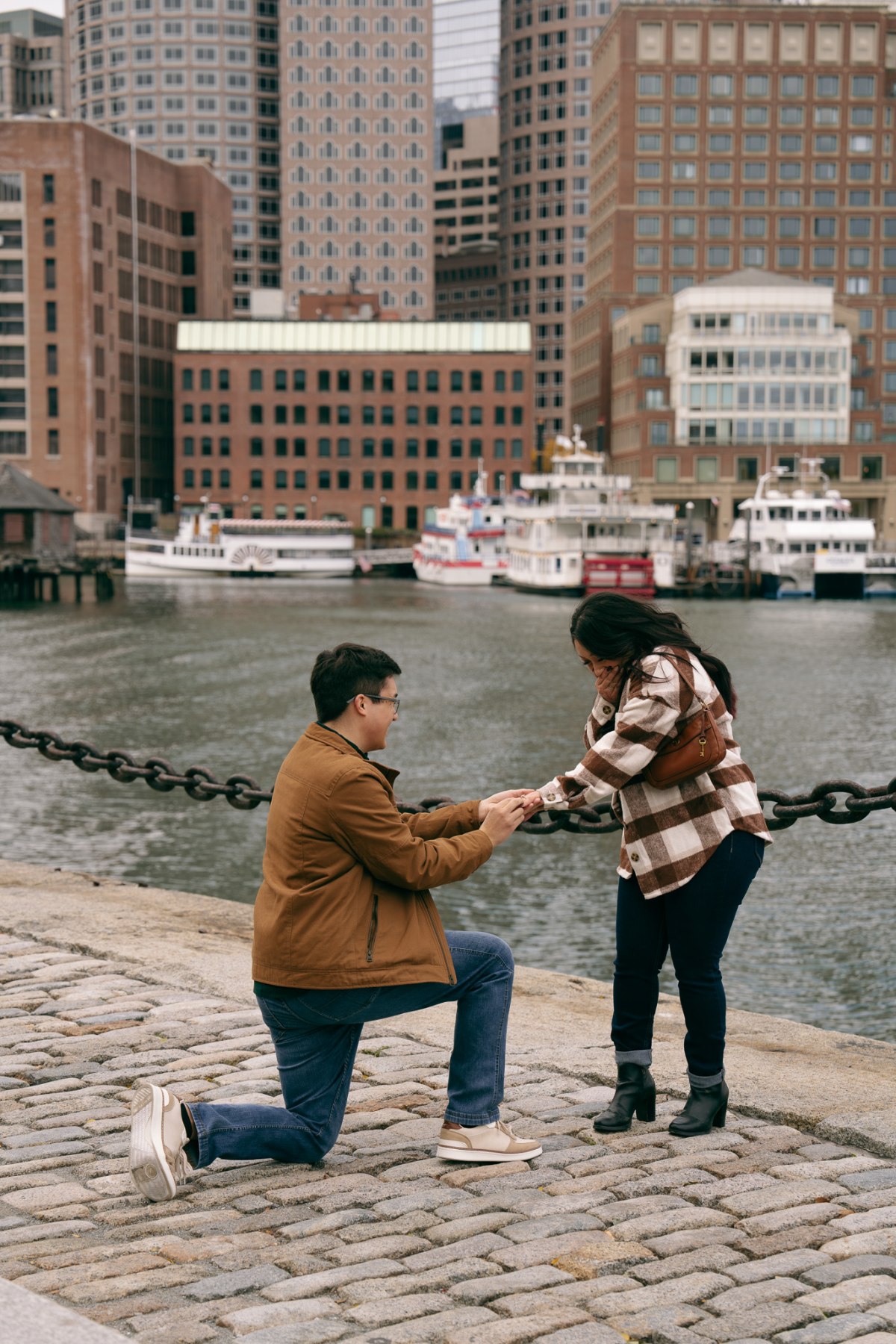 Ethan on one knee proposing to Ariana at Boston Seaport with the harbor behind them