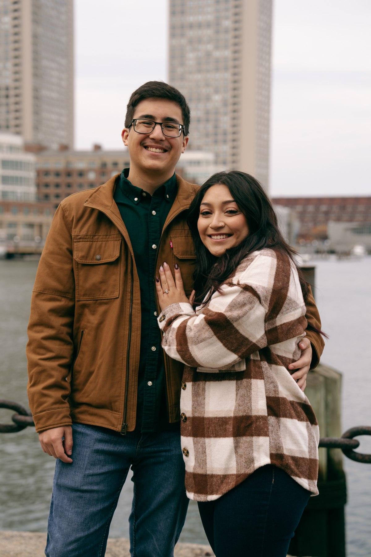 Ethan and Ariana on the curved Fan Pier walkway with the Boston Seaport skyline behind them across the channel