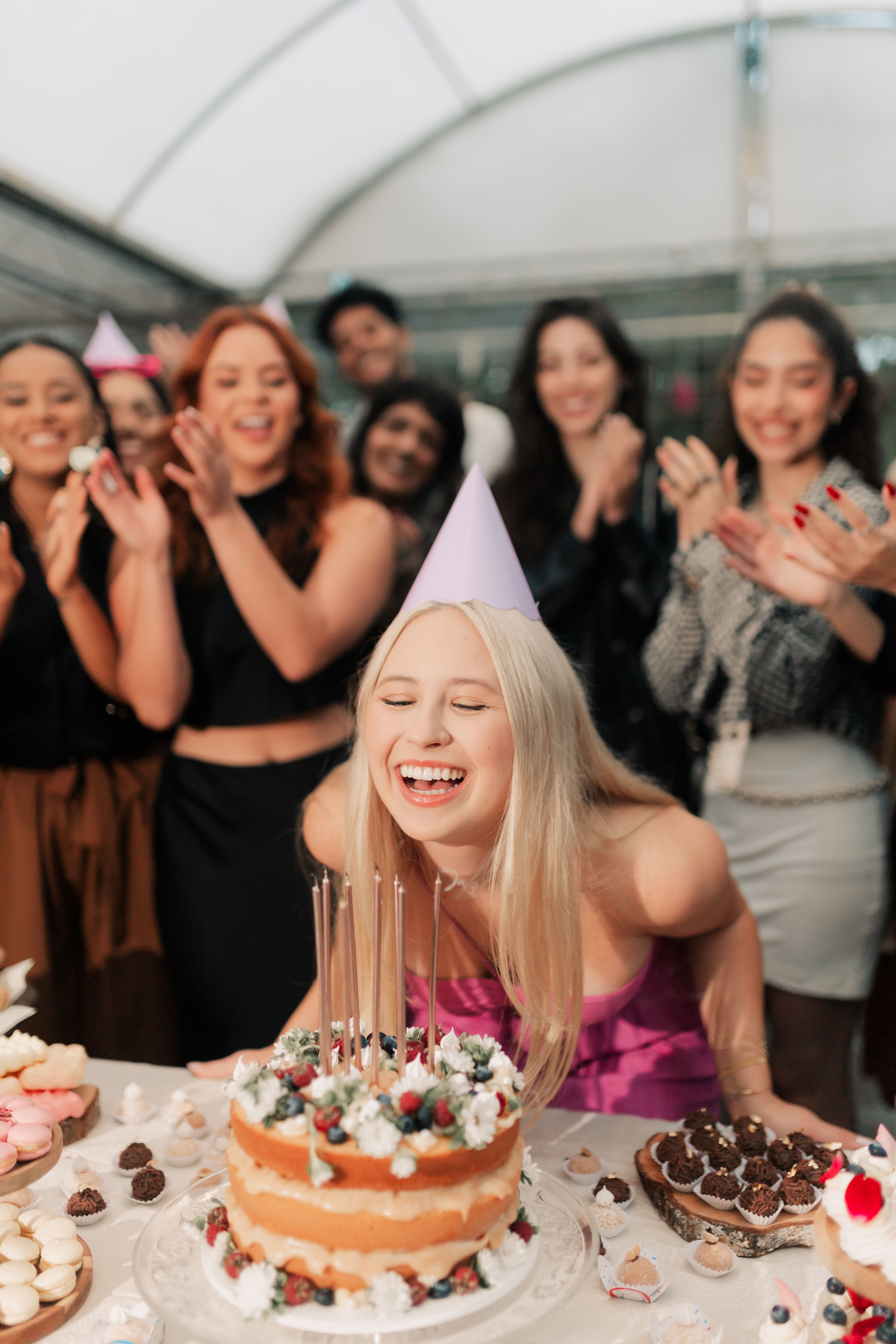 A blonde woman in a lilac party hat laughing joyfully while friends clap in the background — warm, human, editorial celebration moment with strong emotional appeal.
