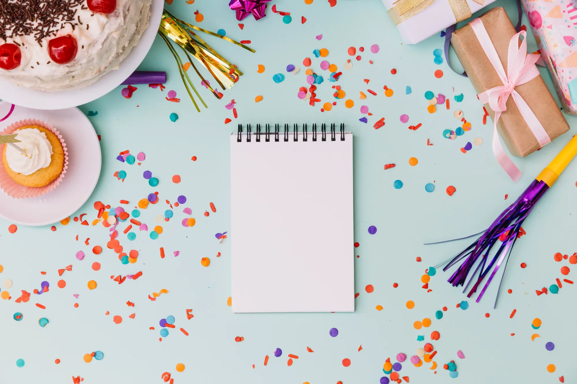 A blank spiral notepad for writing down birthdays, surrounded by cake, wrapped gifts and confetti