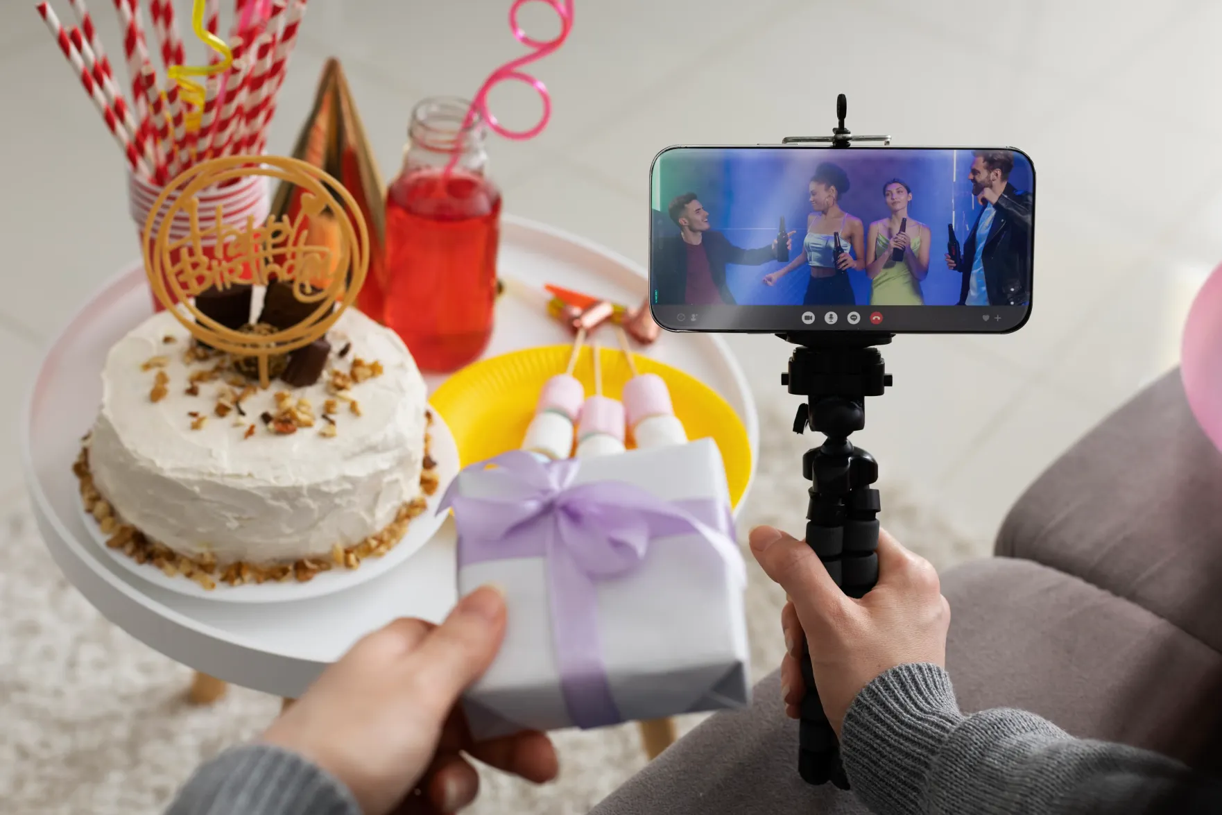 Someone holding up a birthday cake and a wrapped gift to a phone on a tripod during a video call