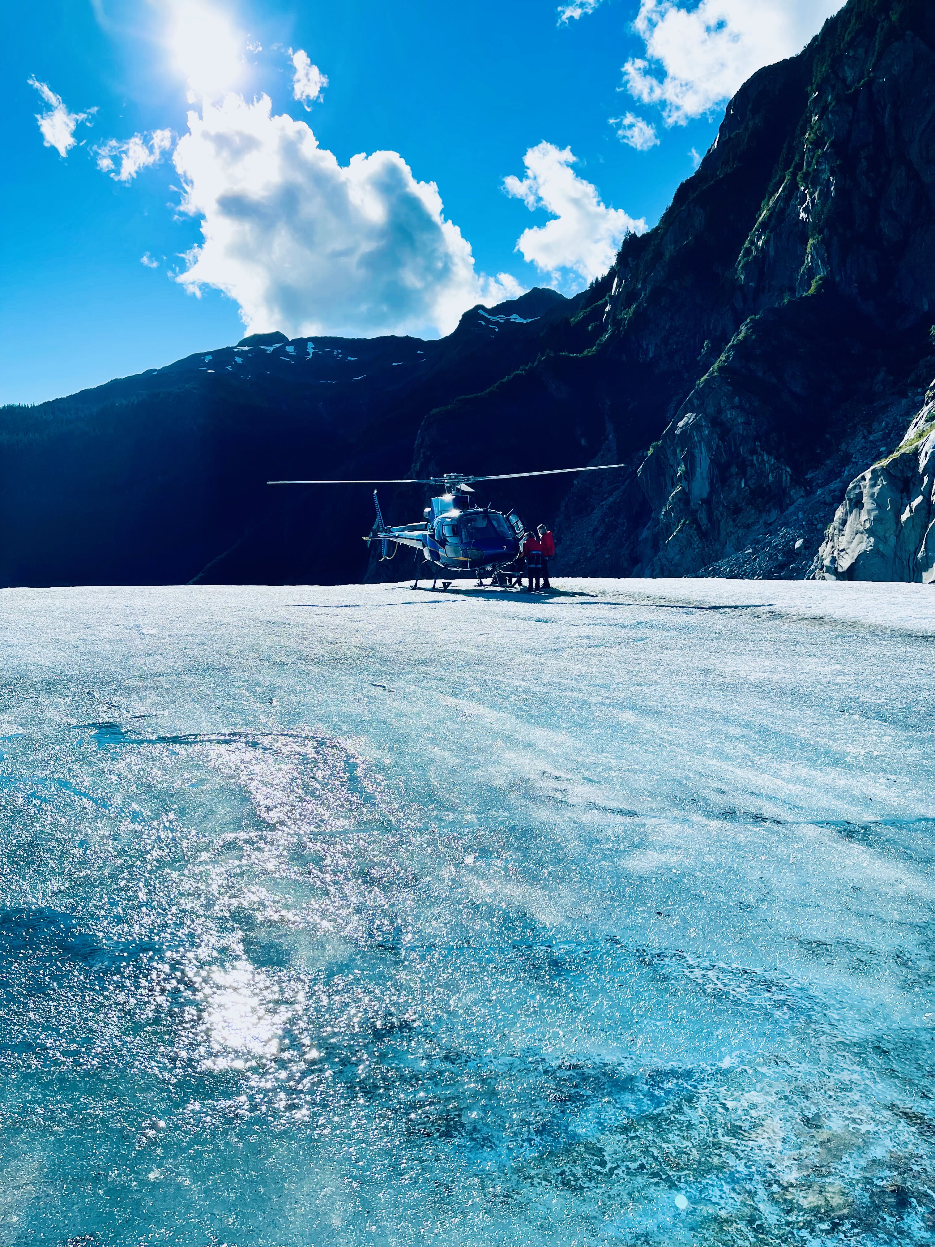 Helicopter on glacier ice with trekker in red beside it and mountains behind
