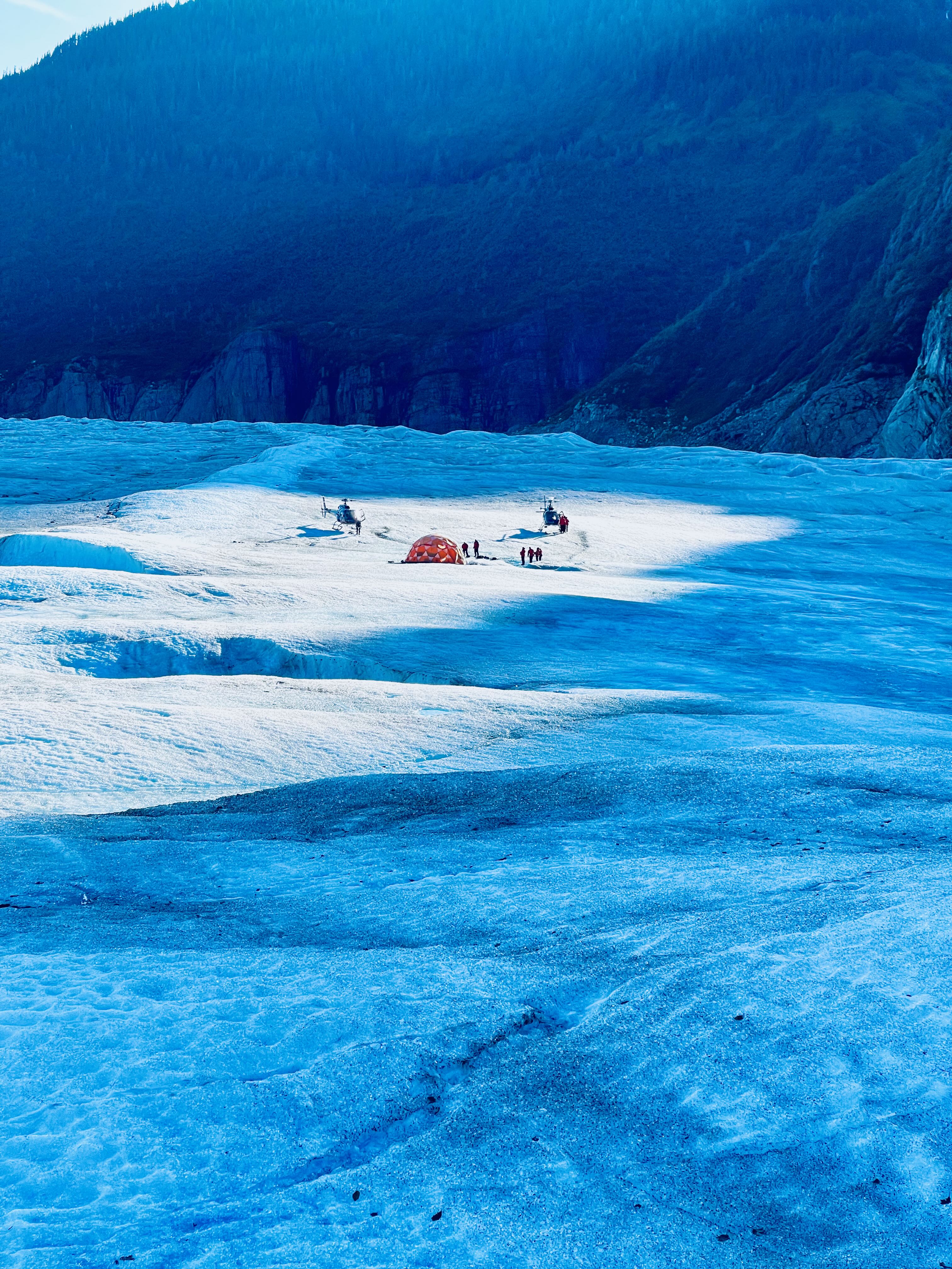 Wide shot of two helicopters and orange basecamp tent tiny against vast Mendenhall ice field