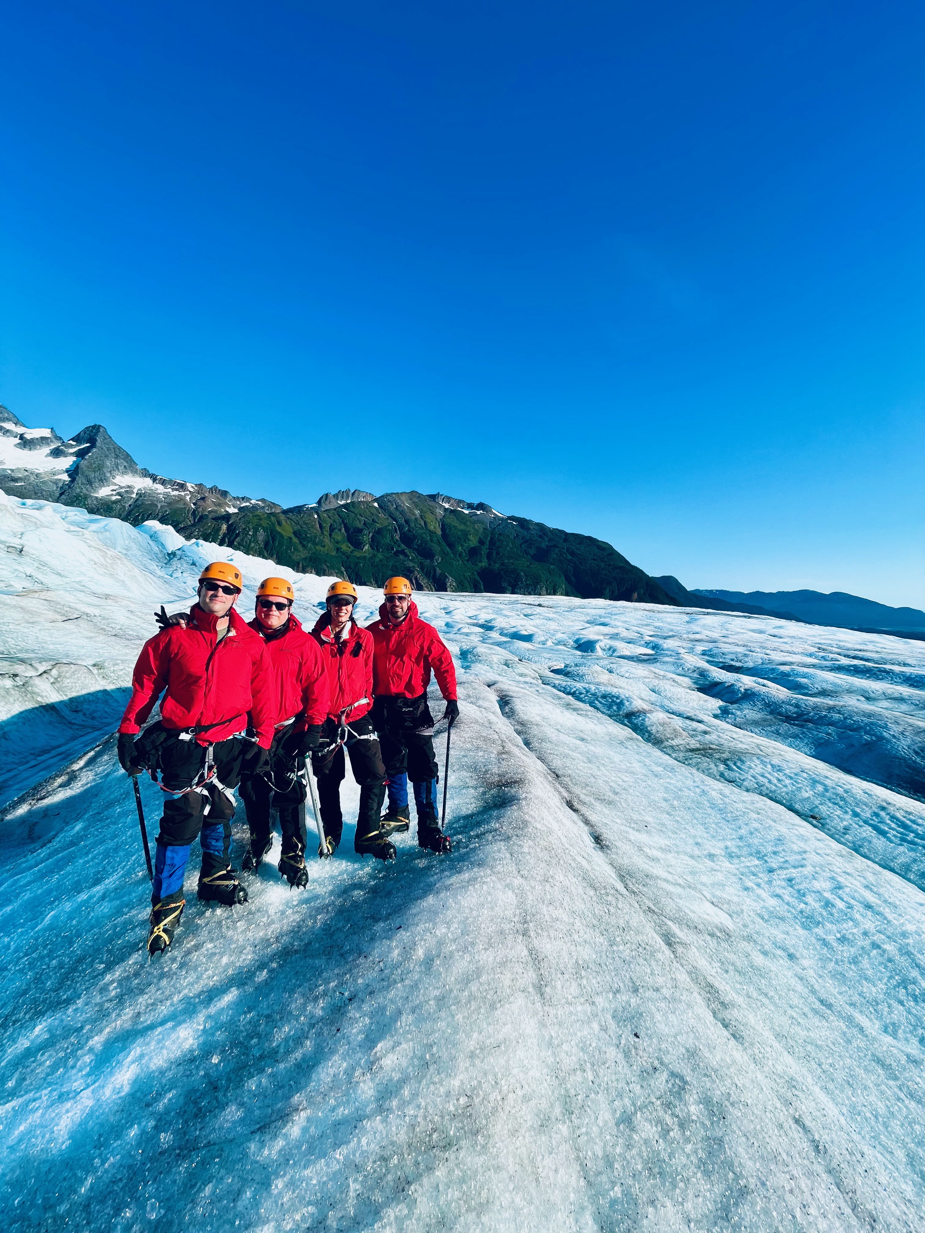 Four crew posed on ice ridge with crampons, ice axes, smiling, mountains behind — the definitive group shot