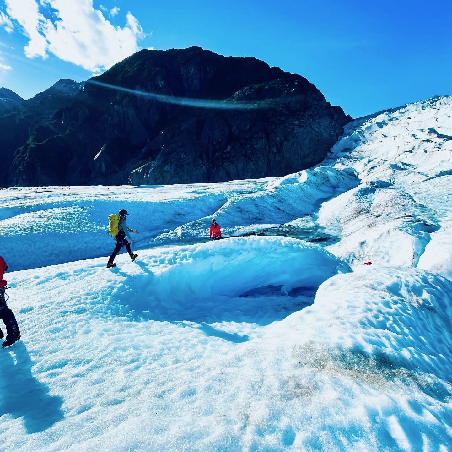 Four trekkers in red jackets on ice ridge with electric blue crevasse below and green mountains behind