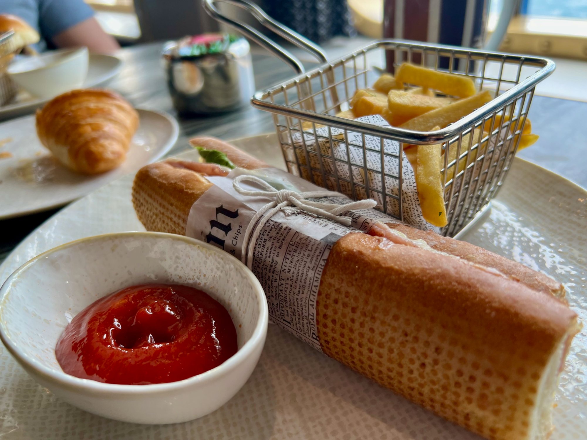 Le Grand Bistro ham baguette sandwich wrapped in newspaper with wire basket of frites