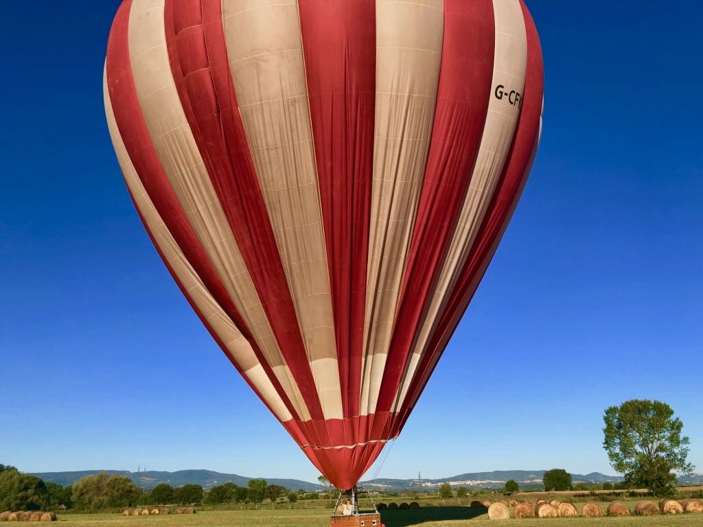 Hot air balloon landing in Tuscan countryside, red and white balloon against blue sky, hay bales in field