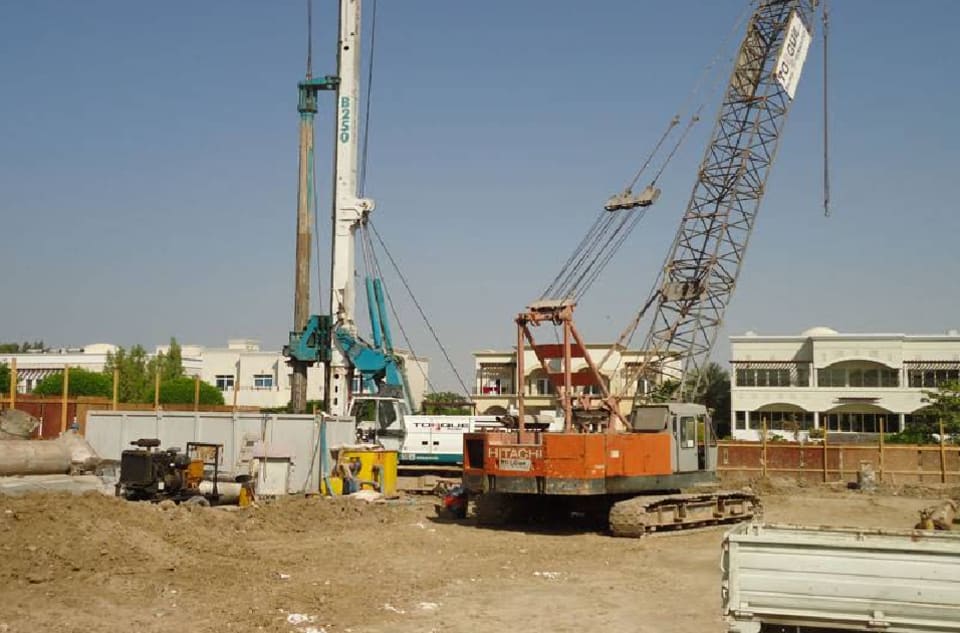 Piling rig and crawler crane mobilised on a villa community site in Dubai, with the Al Jawadri Contracting site board and safety instructions at the gate
