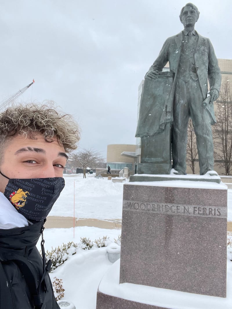 Justin in a Ferris State University mask standing beside the Woodbridge N. Ferris statue in winter