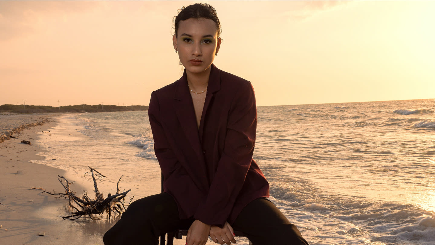 Retrato de una mujer joven sentada en la playa al atardecer. Lleva una chaqueta oscura y mira directamente a la cámara, con el océano y el cielo anaranjado de fondo.