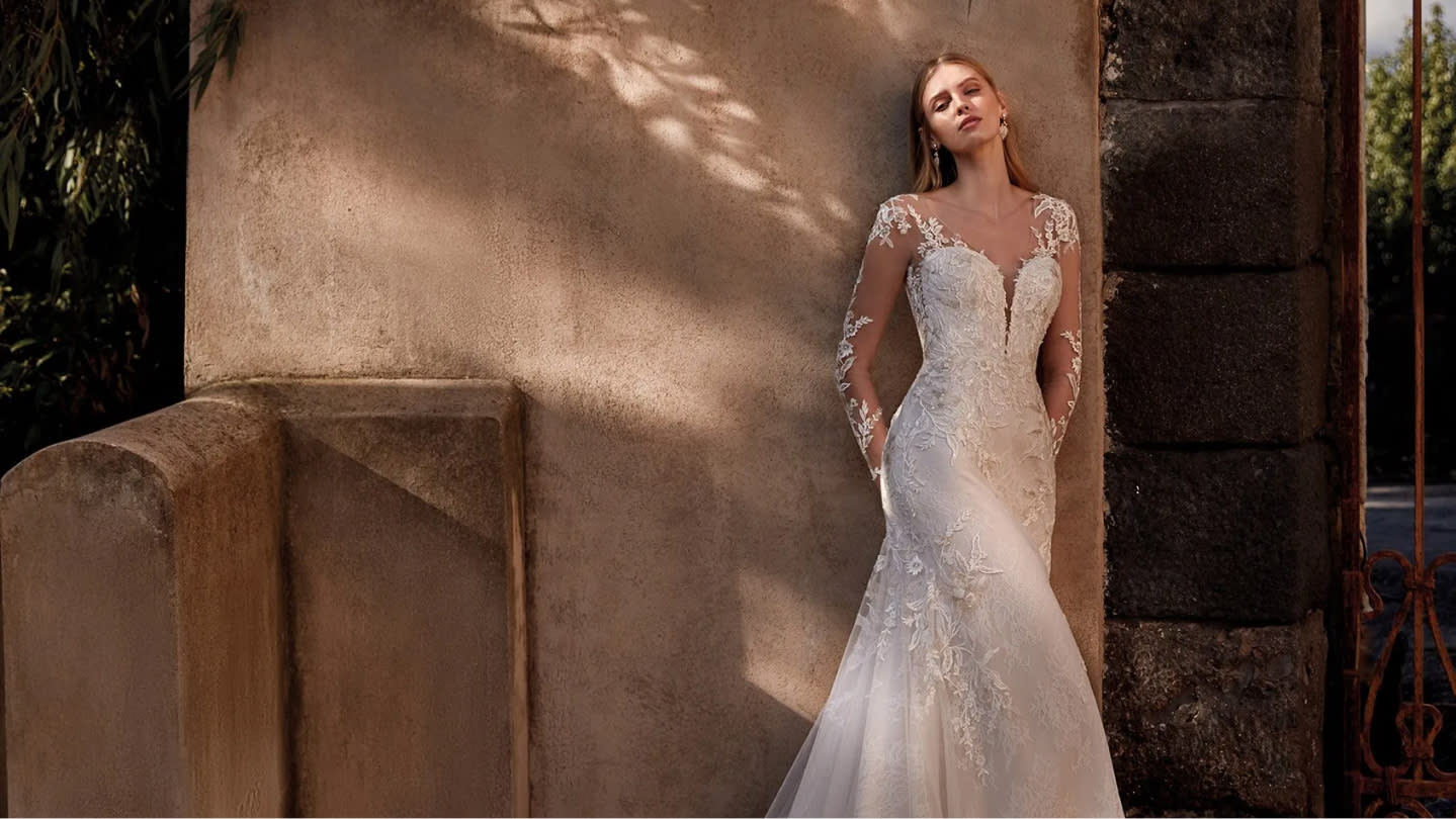 Mujer joven con un vestido de novia de encaje, apoyada contra una pared de piedra antigua, con un entorno natural en el fondo.