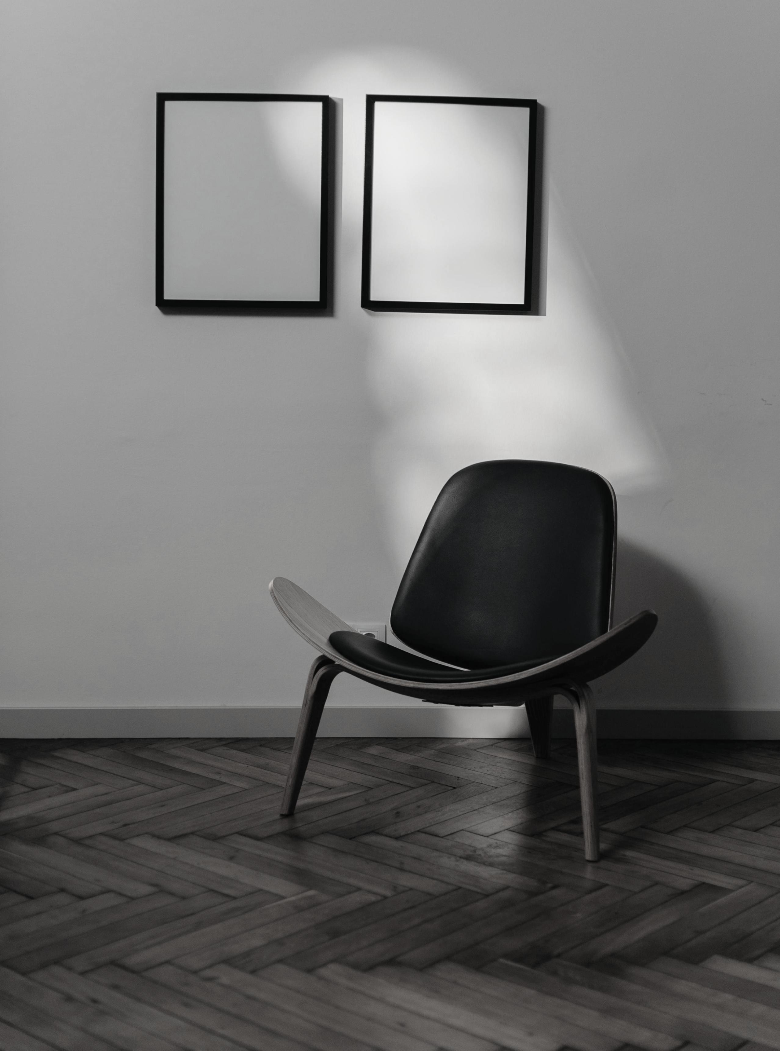 Imagen en blanco y negro de una habitación minimalista con dos cuadros vacíos colgados en la pared y una silla moderna de diseño curvo colocada en el suelo de parqué.