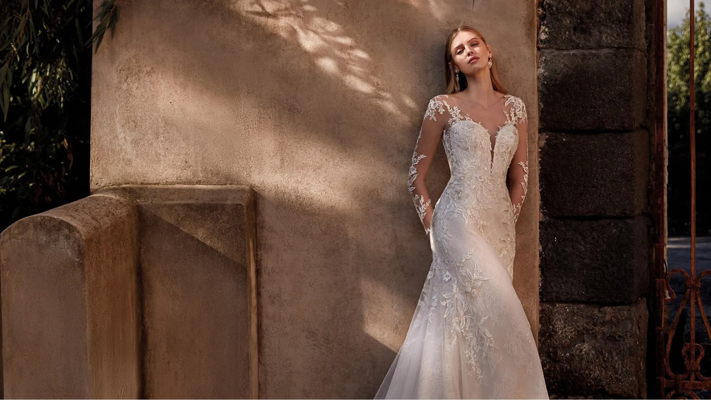 Mujer joven con un vestido de novia de encaje, apoyada contra una pared de piedra antigua, con un entorno natural en el fondo.