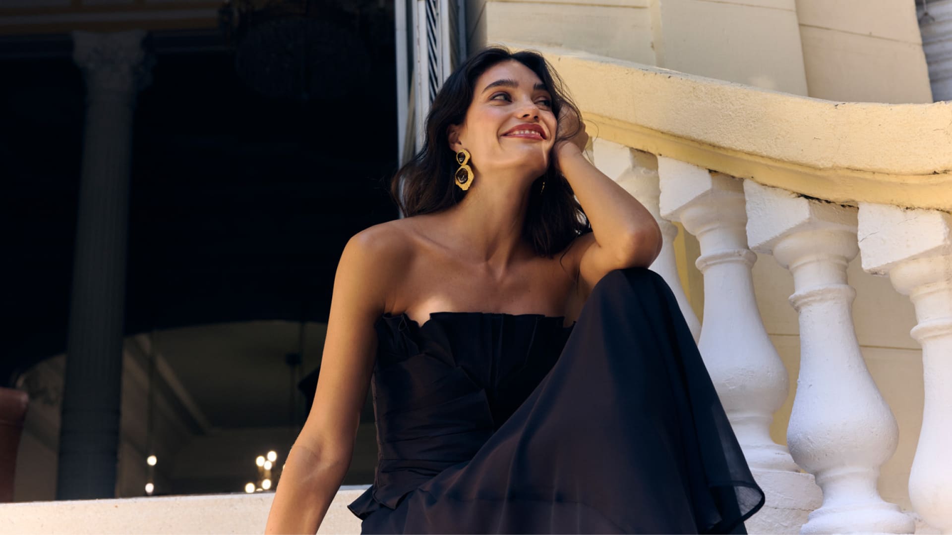 Retrato de una mujer sentada en una terraza, vestida de negro y con pendientes dorados, sonriendo mientras posa junto a una balaustrada blanca iluminada por la luz del atardecer.