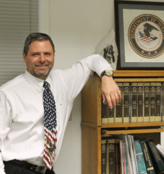 Attorney Risha standing next to a bookshelf resting his left arm on it.