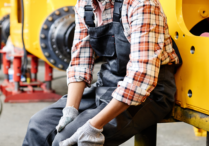 worker of warehouse in coveralls and hardhat touching hurting knee while sitting by industrial machine