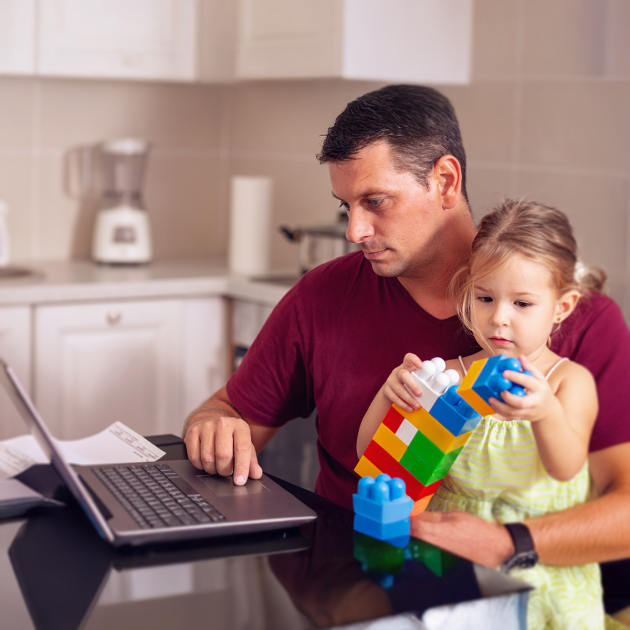 A father working on his laptop while holding his young daughter, who is playing with blocks