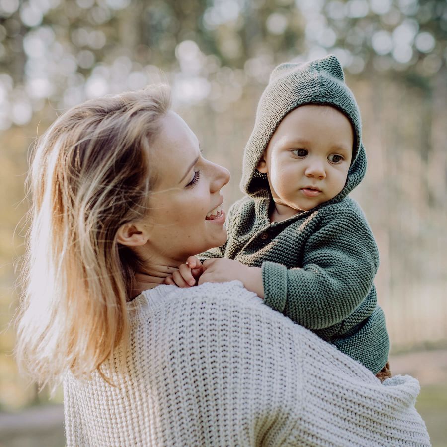 Mother holding her little baby son wearing knitted sweater during walk in nature.