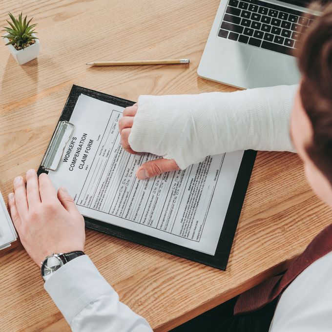 cropped view of worker holding broken arm compensation claim and sitting at table in office, compensation concept